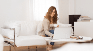 Woman using laptop in cozy living room with white sofa, experiencing year-round comfort from a reverse cycle air conditioner.