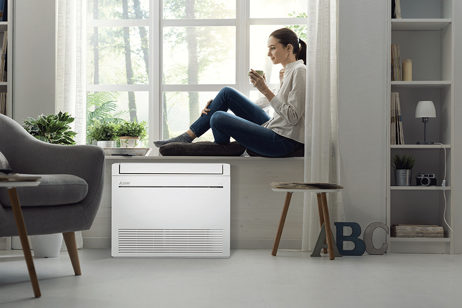 Woman sitting by a window with a hot drink, above a white MFZ-KW HyperCore® floor console heat pump installed below the windowsill in a modern living space.